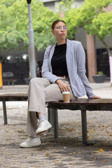 Fototapeta premium African American woman sitting on wooden bench in urban plaza wearing grey blazer holding paper cup
