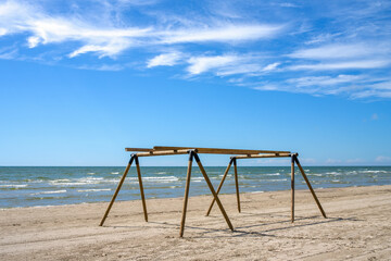 Fototapeta premium Empty wooden canopy frame setup on a sandy beach next to the sea under a bright blue sky with wispy clouds in summer.