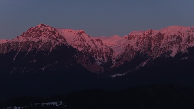 Majestic intense red alpenglow on the majestic snow capped Bucegi Mountains peaks during sunset, uninterrupted tele aerial pan, cinematic 4K