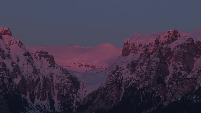 Stunning pink alpenglow on the steep snow capped peaks of Batrana, Gutanu and Omu in the Bucegi Mountains, aerial pan left 168mm super telephoto cinematic 4K