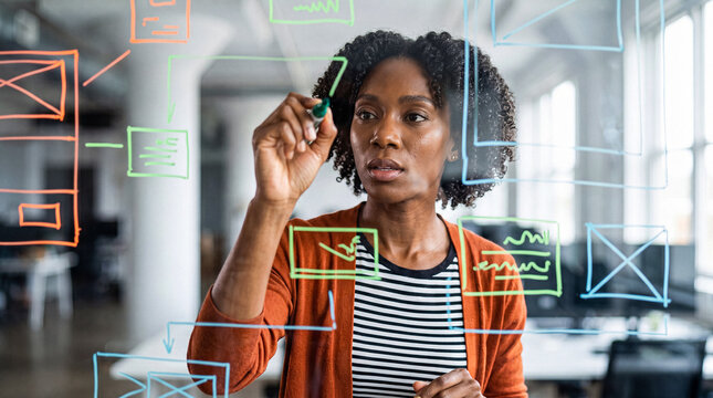 Creative Black woman designing user experience wireframes on a transparent glass board in a modern office space.
