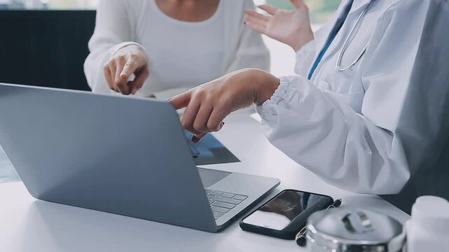 Close-up of a doctor in a white lab coat pointing at a laptop screen while a patient in a white cardigan looks on, with a stethoscope draped around the doctor's neck and a smartphone and medical