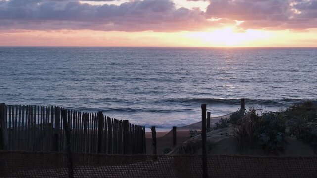 Static shot of a peaceful sunset over the calm ocean waves from the dunes of Labenne-Ocean with wooden fences, Landes, France