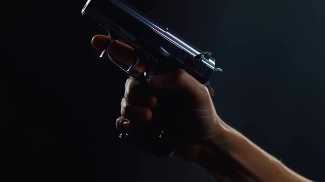 Slow motion tracking shot of hand raising and firing a handgun with recoil seen in dark studio setting with low key rim lighting and visible metal slide movement