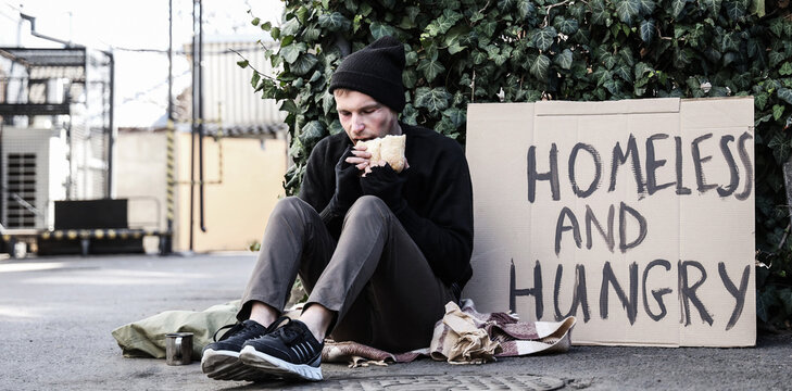 Homeless man with cardboard eating bread on street