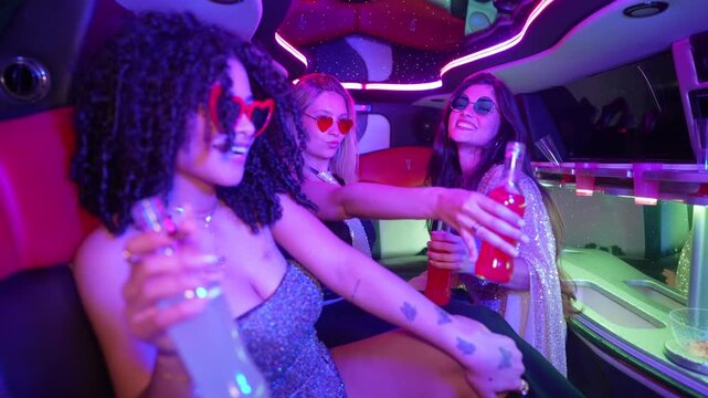 Three happy young women dancing, laughing, and drinking cocktails while riding in a luxury limousine. Stylish friends enjoying a vibrant bachelorette party or prom night with colorful neon lights