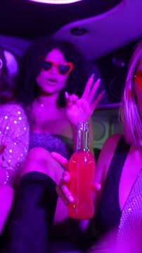 Group of happy young women in stylish party outfits dancing, singing, and drinking inside a luxury limousine with colorful flashing lights during a fun night out on the town