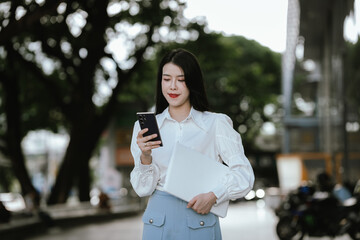 Fototapeta premium Confident young businesswoman holding a smartphone and tablet outdoors in a modern urban setting, representing mobile technology, digital communication, productivity, and professional lifestyle.