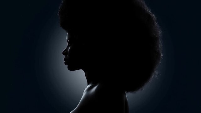 Slow motion profile of woman turning head with afro against dark studio background with subtle halo backlight and soft rim lighting