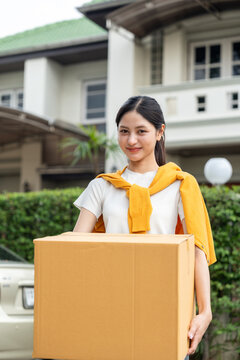 Happy young woman holding moving box and house keys in front of new home. Moving day, real estate purchase, mortgage loan, home ownership and property investment concept.