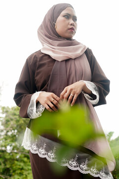 Confident Asian Muslim woman in modest dress looking away behind blurred foreground leaves.