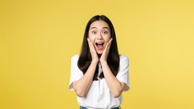 Surprised Girl on Yellow Background: A young Asian girl with a look of pure astonishment, her hands pressed to her cheeks, gasps with wide eyes against a vibrant yellow backdrop.