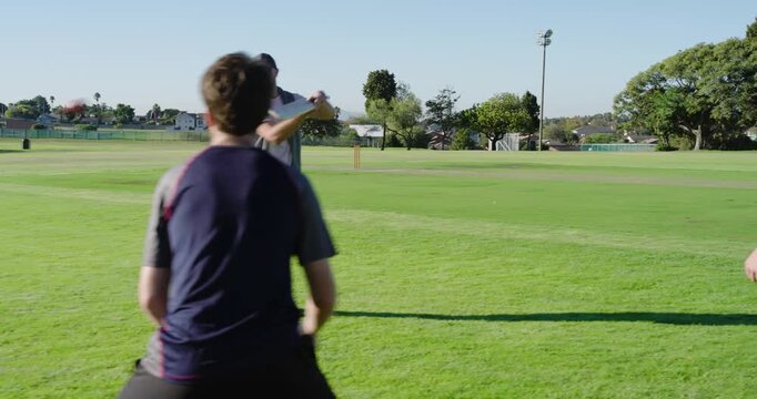 Male coach tapping red ball off bat for male players practicing fielding on cricket field