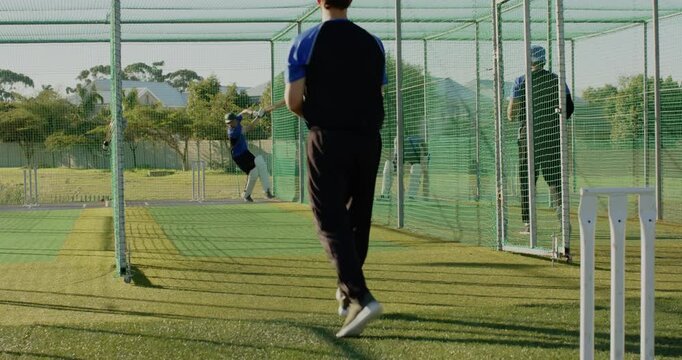 Practicing male teammates swinging bats inside net cage on turf, with stumps, copy space