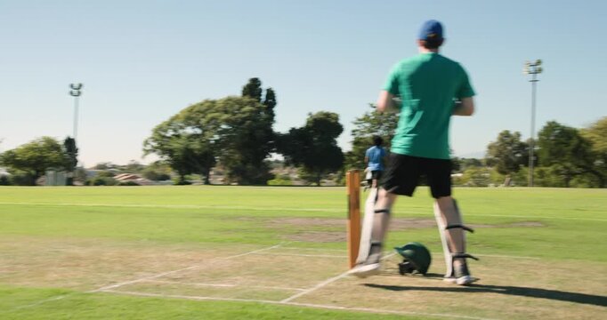 Batsman standing at crease on marked grass pitch, helmet, fielders and floodlight towers