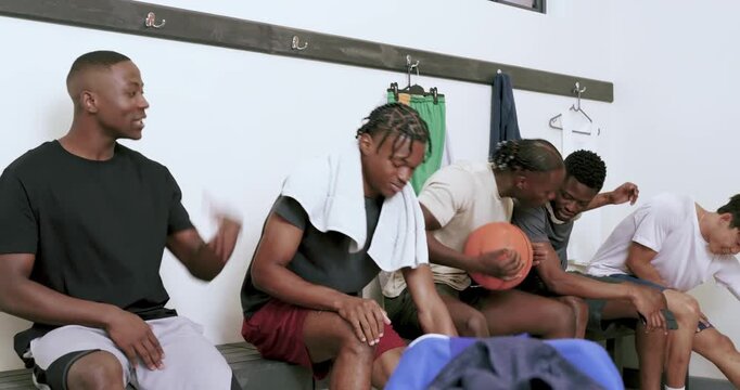 Post-practice, diverse teammates bouncing ball and sharing smartphone on locker room wooden bench