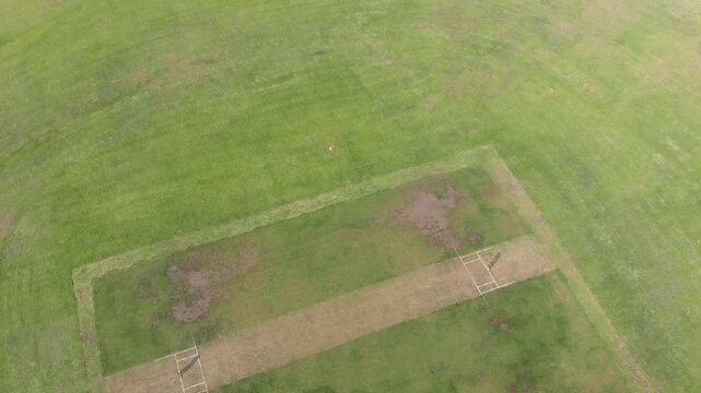 On command drone camera descending, tilting over grassy field revealing cricket pitch and stumps