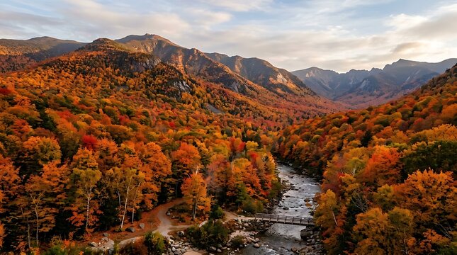 Aerial view of a vibrant autumn landscape with a winding river and bridge through a forest of colorful trees surrounded by mountains under a cloudy sky.