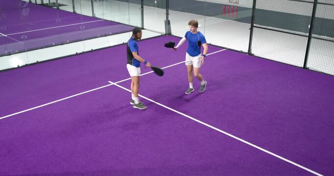 Two male paddle teammates reacting to serve on purple court, exchanging paddle pat and resetting
