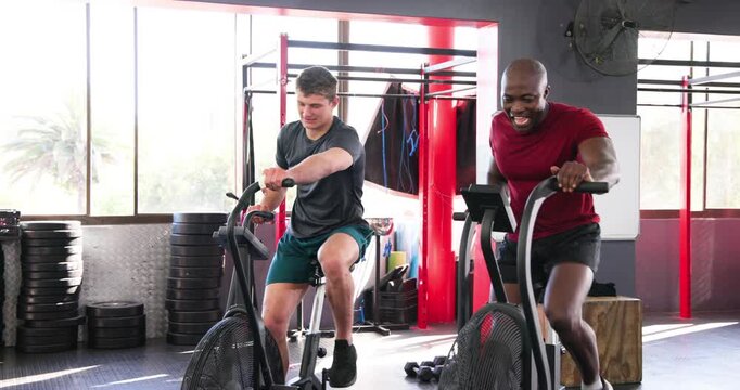 Diverse male duo entering gym and mounting air bikes pedaling for hiit and exchanging nods