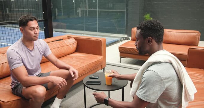 Diverse male friends in lounge exchanging device, discussing, sipping juice, shaking hands