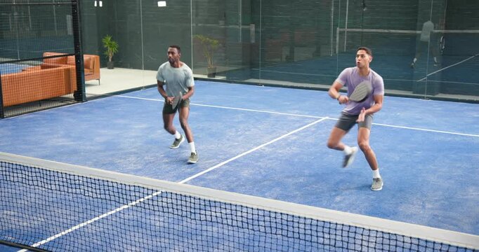 Diverse male partners on padel court responding to ball appearing lunging, swinging paddles