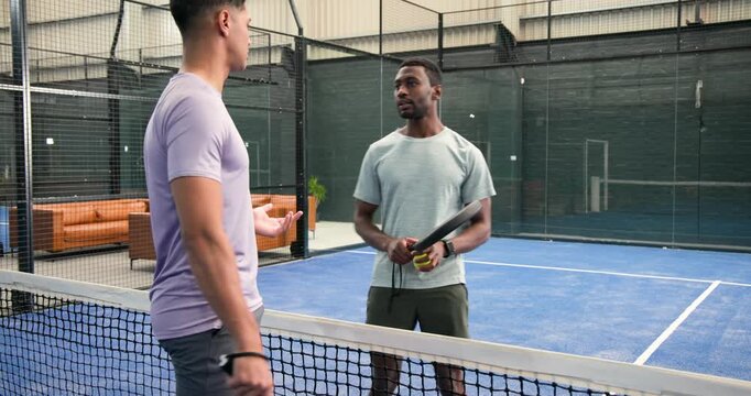 Diverse male friends conversing on padel court by net, fist bumping then rallying with rackets