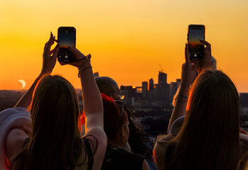 Backlit people, from the top of the Eiffel Tower, capturing a beautiful partial solar eclipse with their smartphones against a golden sunset sky with the Paris skyline and La Défense in the background