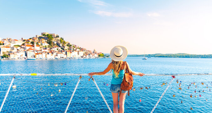 Female tourist with straw hat and backpack enjoying scenic panoramic view of Sibenik old town from a bridge with love padlocks, authentic summer travel lifestyle in Croatia
