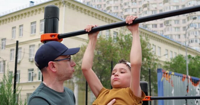 Father helps son with pull-up exercise at playground. Child shows determination and focus while training. Outdoor fitness promotes healthy lifestyle in urban environment. Parental training.