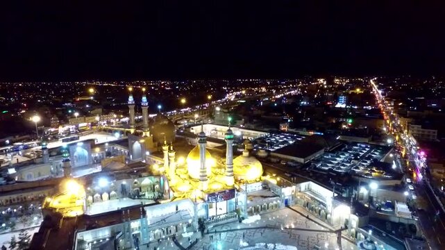 High drone overview of the beautifully lit central courtyards and golden minarets of the holy shrine of Lady Masoumeh surrounded by a glowing urban cityscape under dark evening skies.