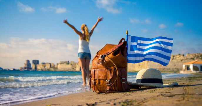 Leather backpack with greek flag and straw hat on the beach, female tourist raising arms in blurred background, authentic travel freedom concept in Greece