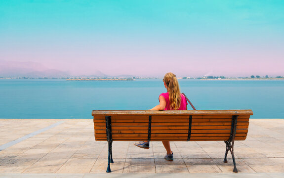 back view of woman sitting on bench looking at calm sea during pastel sunset