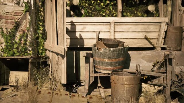 Wooden buckets sit on a porch surrounded by greenery, with a rustic backdrop. The sun casts shadows, enhancing the peaceful, vintage atmosphere of this outdoor space.