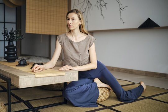 Focused woman sits on a mat with her legs crossed at a low table. The room has minimal decor with natural light coming in and a calm atmosphere