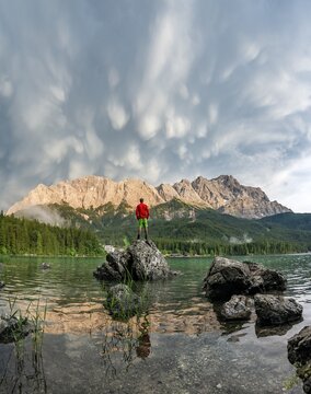Young man standing on a rock on the shore, Eibsee lake in front of Zugspitze massif with Zugspitze, sunset, dramatic Mammaten clouds, Wetterstein range, near Grainau, Upper Bavaria, Bavaria, Germany