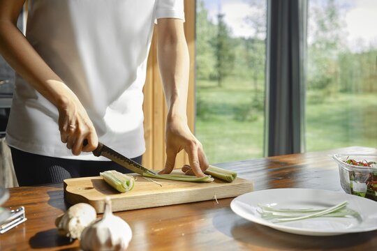 Unrecognizable woman skillfully slices green leek on a wooden cutting board in a bright kitchen with outdoor views