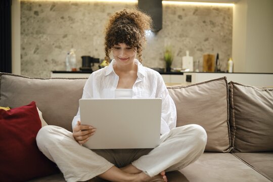 A young woman sits on a couch, laptop on her lap, focused on her work or leisure activity. She is dressed casually, enjoying a cozy atmosphere in her modern living room during the day