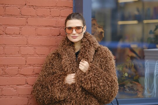 A woman poses confidently in a warm, brown, fluffy coat and oversized sunglasses. She stands against a red brick wall, showcasing a modern urban style in a vibrant setting