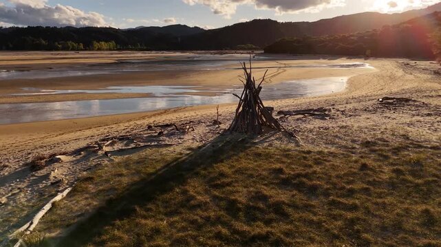 Driftwood firepit structure on vast tidal sandflat with shallow reflective pools, estuary and bush-covered hills glowing under golden sunset light in Abel Tasman National Park, New Zealand.Aerial view