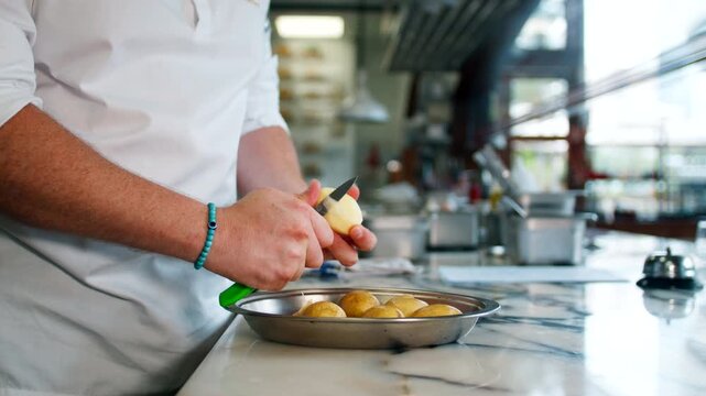 Side view of a professional male chef peeling raw potatoes with a hand peeler tool over a steel workstation countertop, preparing fresh ingredients in a restaurant kitchen