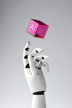 A robotic white hand reaches for a levitating purple transparent cube with "AI" inscription against a light gray background. Concept of artificial intelligence, futuristic technology and innovation.