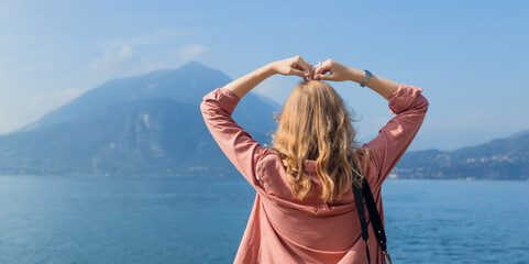 young blonde woman from behind stands by a mountain lake and creates a heart symbol with her hands, framing the beautiful nature scenery. Concept of love for travel, freedom, wanderlust, and summer © Shi 
