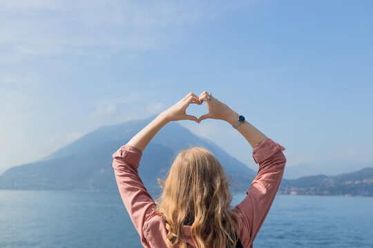 young blonde woman from behind stands by a mountain lake and creates a heart symbol with her hands, framing the beautiful nature scenery. Concept of love for travel, freedom, wanderlust, and summer