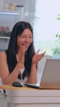 Asian woman using laptop with smile at home desk, happy remote work on computer indoor workspace, bright office interior with casual technology success scene Vertical