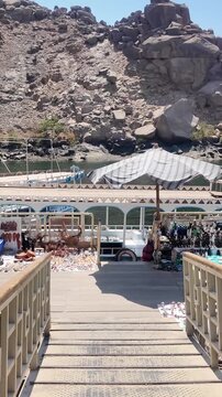 First person view walking down a ramp towards a floating market on the Nile River. Souvenirs for sale are displayed on the dock in philae
