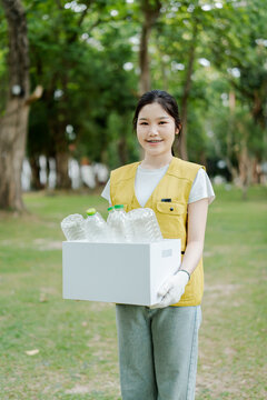 Young asian woman volunteering for environmental protection, holding a box filled with plastic bottles for recycling in a green park, promoting sustainability