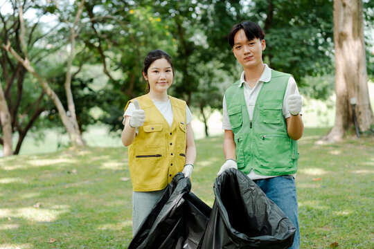 Young asian man and woman volunteers wearing vests, holding trash bags, giving thumbs up for environmental cleanup and community recycling in a green park