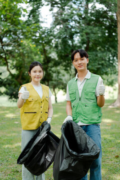 Young asian park clean up volunteers wearing vests and gloves, holding trash bags, giving thumbs up, promoting environmental responsibility and eco-friendly community service