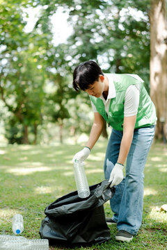 Young asian volunteer collecting plastic bottles into a trash bag, actively participating in a park cleanup to promote recycling and environmental sustainability